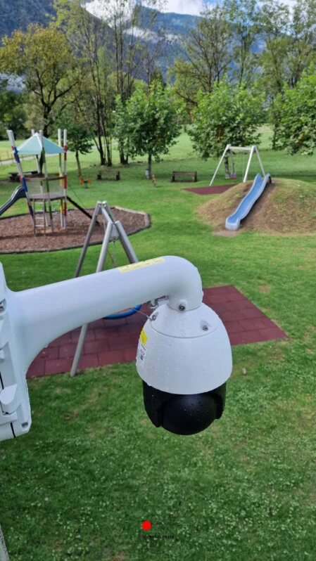 Telecamera PTZ di Hikvision installata in area giochi pubblica all’aperto Telecamera di videosorveglianza PTZ montata vicino a un parco giochi con altalene e scivoli, immerso nel verde con sfondo montano
