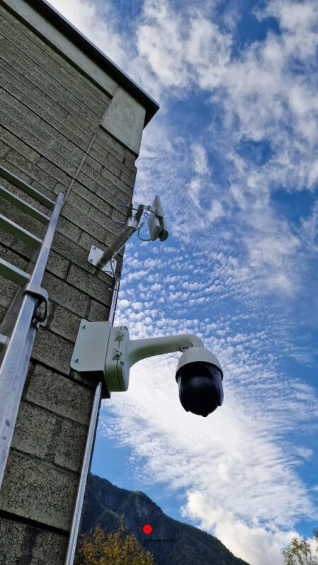 Telecamere di videosorveglianza esterne installate su edificio in montagna Telecamere di videosorveglianza PTZ montate su parete esterna in cemento con sfondo montuoso e cielo nuvoloso
