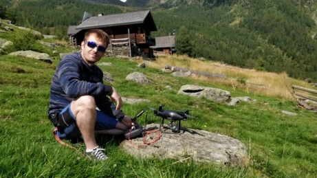 Operatore drone in montagna per riprese aeree panoramiche Pilota di drone seduto su un prato alpino pronto al decollo, con controller e drone posati su roccia, sullo sfondo una baita in legno tra i monti